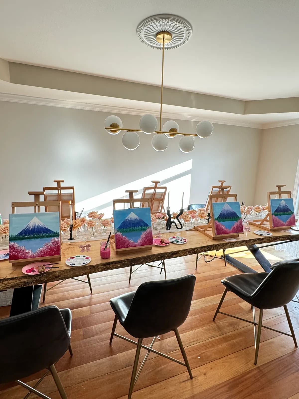 Easels, palettes, and blush florals set along the table under a brass chandelier at a Mind & Body Retreats art workshop