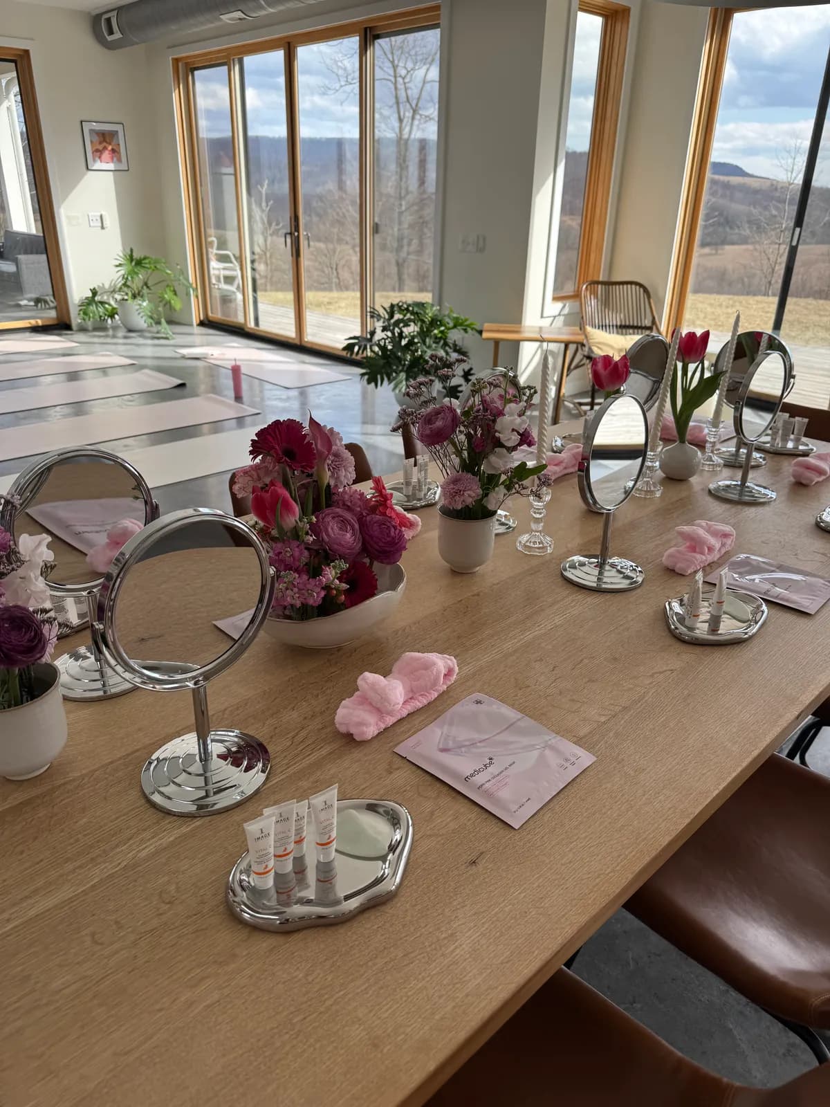 Long workshop table set with vanity mirrors, silver trays, and pink florals in front of floor-to-ceiling windows at a Mind & Body Retreats beauty workshop